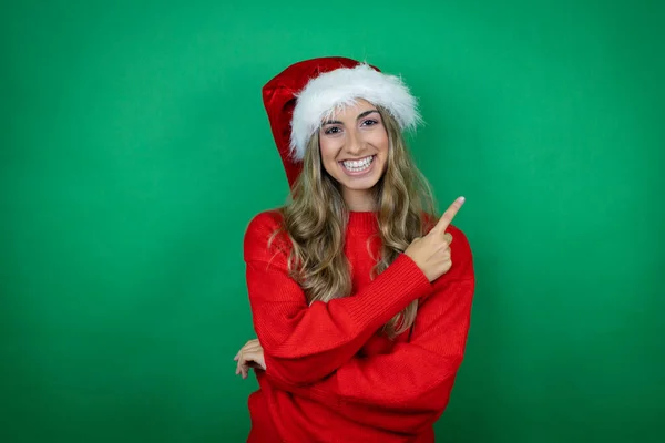 年轻美丽的女孩戴着圣诞圣诞圣诞礼帽 带着礼物 带着孤独的绿色背景微笑 手指手画脚地指向旁边 — 图库照片