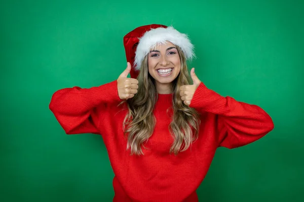 年轻美丽的女孩戴着圣诞圣诞圣诞礼帽 带着礼物 带着孤零零的绿色背景 微笑着 用她的大拇指做着 的手势 — 图库照片