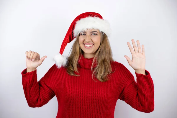 年轻漂亮的金发女人穿着红色的休闲装 头戴一顶白色背景的圣诞帽 带着自信和快乐的笑容 用手指指了指六号 — 图库照片