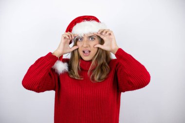 Genç, sarışın, kırmızı bir süveter ve beyaz arka planda bir Noel şapkası takmış parmaklarıyla gözlerini açmaya çalışan, uykulu ve yorgun bir kadın.