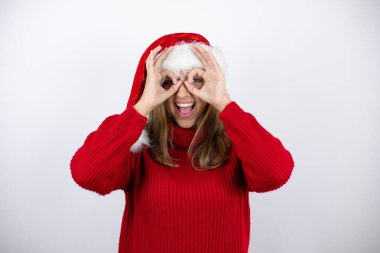 Kırmızı bir süveter ve beyaz arka planda bir Noel şapkası giyen güzel, sarışın bir kadın gülümseyen yüz ve parmaklarından bakan gözlerle şok olmuş bir jest yapıyor.