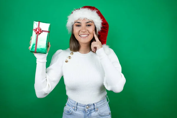 年轻美丽的女人戴着圣诞礼帽 身穿白色背景的白色毛衣 满面春风 自信地微笑着 用手指 牙齿和嘴指指点点着 — 图库照片