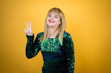 Sarı arka planda süslü bir elbise giyen güzel sarışın kadın iki numarayı gösterirken kendinden emin ve mutlu bir şekilde gülümsüyor.