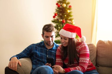 Mutlu ve kendinden emin gülümseyen genç bir çift. Kanepede oturup evdeki Noel ağacının etrafında telefon kullanmak.