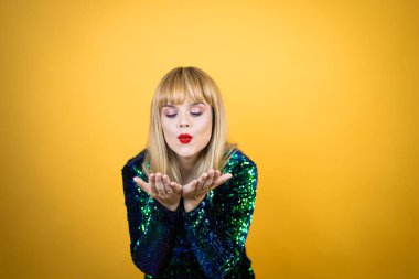 Sarı arka planda süslü bir elbise giyen güzel sarışın kadın avuçlarını birbirine uzatarak gülümsüyor ya da el hareketi yapıyor. Tutun ve koruyun