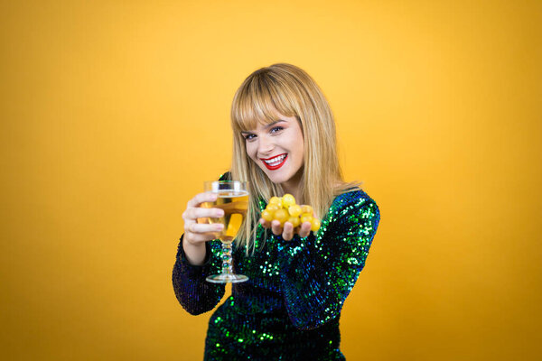 Beautiful blonde woman wearing a fancy dress over yellow background holding a glass of champagne and grapes. New Years Eve celebration