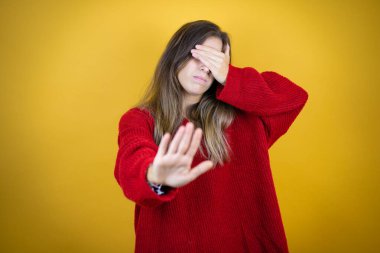 İzole edilmiş sarı arka planda kırmızı süveter giyen genç ve güzel bir kadın ellerle gözlerini kapatıyor ve üzgün ve korku ifadeleriyle dur hareketi yapıyor. Utanç verici ve olumsuz bir kavram..