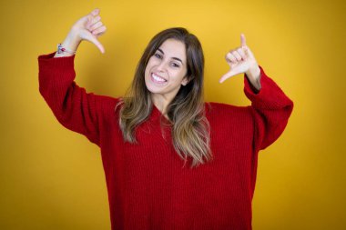 İzole edilmiş sarı arka planda kırmızı süveter giyen genç ve güzel bir kadın. Kendine güvenen, yüzünde gülümseme olan, kendini gururlu ve mutlu gösteren bir kadın..