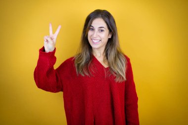 İzole edilmiş sarı arka planda kırmızı süveter giyen genç ve güzel bir kadın iki numaralı parmaklarıyla işaret ederken kendinden emin ve mutlu bir şekilde gülümsüyor.