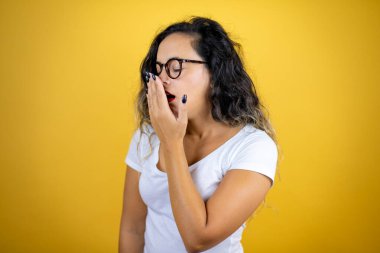 İzole edilmiş sarı arka plan üzerine gündelik beyaz tişört giyen genç ve güzel bir kadın ağzıyla esnemekten sıkılmış. Huzursuz ve uykulu..