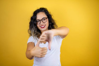 İzole edilmiş sarı arka plan üzerinde günlük beyaz tişört giyen genç, güzel bir kadın baş parmaklarını yukarı aşağı hareket ettiriyor, anlaşmazlık ve anlaşma ifadesi takınıyor. Çılgın çatışma