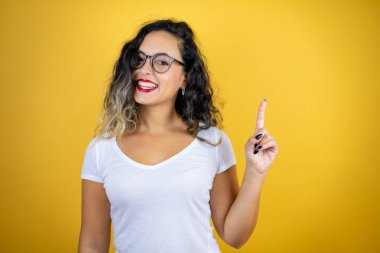 İzole edilmiş sarı arka plan üzerine günlük beyaz tişört giyen genç ve güzel bir kadın kendini beğenmiş ve mutlu bir şekilde gülümserken bir numaralı parmaklarıyla işaret ediyor.