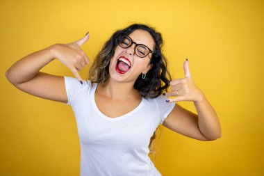 İzole edilmiş sarı arka plan üzerine günlük beyaz tişört giyen genç ve güzel bir kadın çılgın bir ifadeyle elleri havada rock sembolü yapıyor.