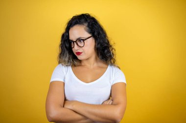 Genç, güzel bir kadın, izole edilmiş sarı arka plan üzerine günlük beyaz tişört giyip kollarını kavuşturmaktan sıkılmış ve yorgun görünüyor.