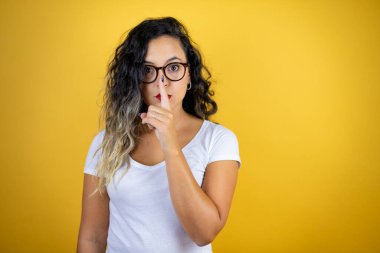 Genç, güzel bir kadın izole edilmiş sarı arka plan üzerine günlük beyaz tişört giyip sessiz olmayı diliyor. Sessizlik ve gizli konsept.