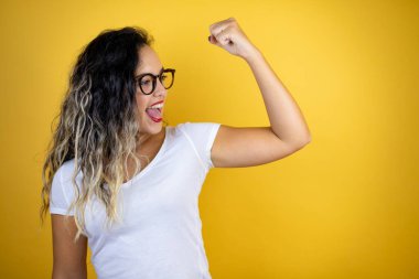 Genç, güzel bir kadın izole edilmiş sarı arka planda günlük beyaz tişört giyiyor. Kollarında gururla gülümseyen kaslar var.