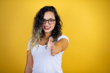 İzole edilmiş sarı arka planda günlük beyaz tişört giyen genç ve güzel bir kadın selamlama ve hoş geldin demek için gülümseyerek tokalaşıyor.