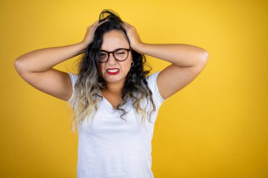 Genç, güzel bir kadın izole edilmiş sarı arka plan üzerine günlük beyaz tişört giyiyor. Baş ağrısı, çaresiz ve stresli. Çünkü elleri başının üstünde acı ve migren var.