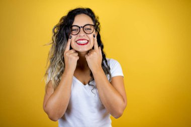 İzole edilmiş sarı arka planda günlük beyaz tişört giyen genç ve güzel bir kadın gülümsüyor ve parmaklarıyla ve ağzıyla işaret ediyor.