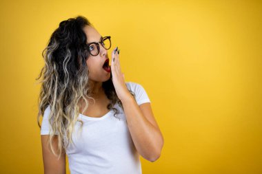 Genç, güzel bir kadın, izole edilmiş sarı arka plan üzerine gündelik beyaz tişört giyiyordu. Eli ağzının üzerindeydi ve şaşırmıştı.