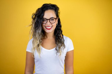 İzole edilmiş sarı arka plan üzerine günlük beyaz tişört giyen genç ve güzel bir kadın. Mutlu bir yüzü olan, kendine güvenen bir gülümsemeyle dişlerini gösteren.