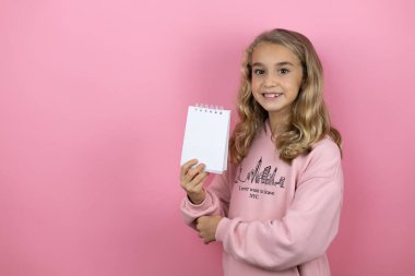 Genç, güzel bir kız çocuğu pembe arka planda duruyor gülümsüyor ve elinde boş bir defter gösteriyor.