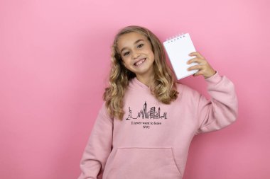 Genç, güzel bir kız çocuğu pembe arka planda duruyor gülümsüyor ve elinde boş bir defter gösteriyor.