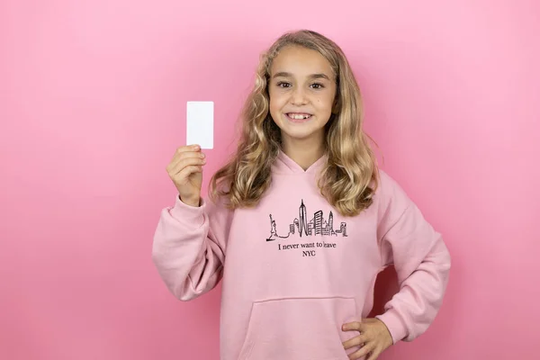 Güzel bir kız çocuğu pembe arka planda duruyor gülümsüyor ve elinde beyaz bir kart tutuyor.