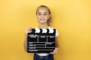 Sarı arka planda duran genç ve güzel bir kız çocuğu elinde bir klaket tutarak çok mutlu.