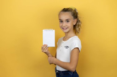 Sarı arka planda duran, gülümseyen ve elinde boş bir not defteri olan güzel bir kız çocuğu.