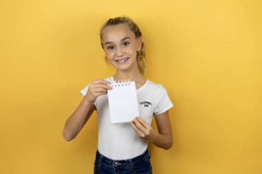 Sarı arka planda duran, gülümseyen ve elinde boş bir not defteri olan güzel bir kız çocuğu.