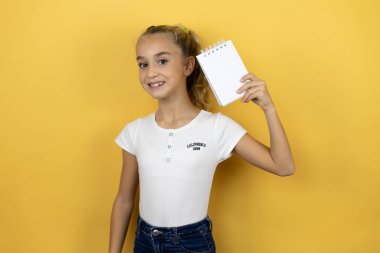 Sarı arka planda duran, gülümseyen ve elinde boş bir not defteri olan güzel bir kız çocuğu.