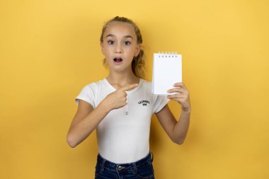 Sarı arka planda duran, gülümseyen ve elinde boş bir not defteri olan güzel bir kız çocuğu.