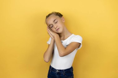 Güzel bir kız çocuğu, izole edilmiş sarı arka planda ayakta uyur, yorgun düşler kurar, elleriyle poz verir ve gözleri kapalı gülümserdi..