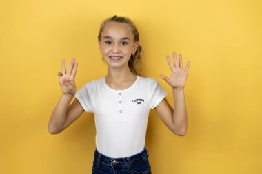 İzole edilmiş sarı arka planda duran genç ve güzel bir kız çocuğu sekiz numaralı parmaklarıyla işaret ederken kendinden emin ve mutlu bir şekilde gülümsüyor.