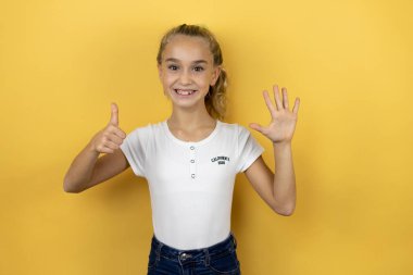 Genç, güzel bir kız çocuğu, izole edilmiş sarı arka planda duruyor ve altı numaralı parmaklarıyla işaret ederken kendinden emin ve mutlu bir şekilde gülümsüyor.