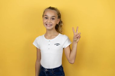 Güzel bir kız çocuğu, izole edilmiş sarı arka planda duruyor ve iki numaralı parmaklarıyla işaret ederken kendinden emin ve mutlu bir şekilde gülümsüyor.
