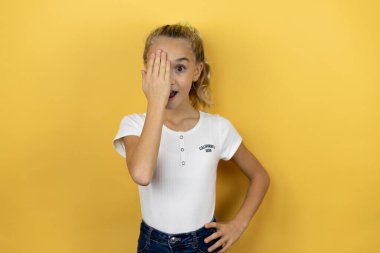 Güzel bir kız çocuğu, izole edilmiş sarı arka planda duruyor. Bir gözünü eliyle kapatıyor. Yüzünde kendine güvenen bir gülümseme ve sürpriz bir duygu var..
