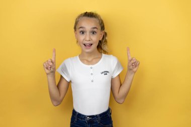 Güzel bir kız çocuğu, izole edilmiş sarı arka planda duruyor. Kameraya bakıyor, parmaklarıyla işaret ediyor ve kollarını kaldırıyor.