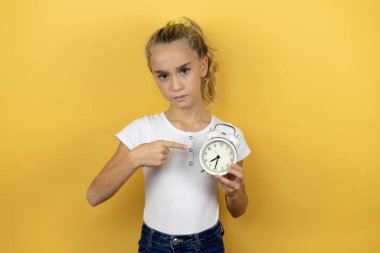 İzole edilmiş sarı arka planda duran genç ve güzel bir kız çocuğu ciddi bir şekilde saati tutuyor ve işaret ediyor.