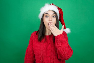 Noel Baba şapkası takan genç ve güzel bir kadın izole edilmiş yeşil arka planda ağzını kapatırken şaşırdı.