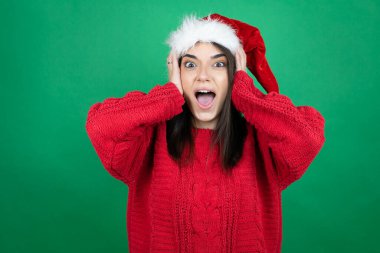 Noel Baba şapkası takan genç, güzel bir kadın yeşil arka planda çılgın ve korkmuş, korkmuş ve şoktan korkmuş.
