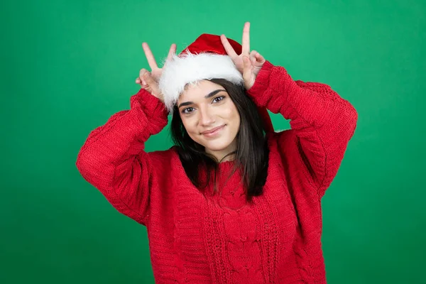若いです美しい女性身に着けていますクリスマスサンタ帽子隔離された緑の背景上にウサギの耳として頭の上に指で面白いとクレイジーをポーズ 明るい笑顔 — ストック写真