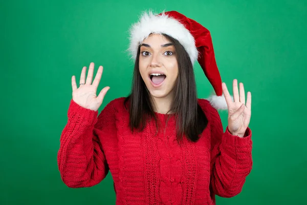 若いです美しいです女性身に着けていますクリスマスサンタ帽子上の隔離された緑の背景ショーと指差す指番号9ながら笑顔自信と幸せ — ストック写真