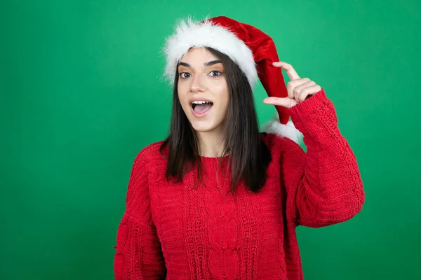 孤立した緑の背景笑顔の上にクリスマスサンタの帽子を身に着けている若い美しい女性と指で小さなサイズの看板を行う手で自信を持ってジェスチャー 概念の測定 — ストック写真