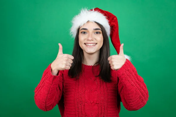 若いです美しいです女性身に着けていますクリスマスサンタ帽子上の隔離された緑の背景笑顔と行いますザOk信号とともに彼女の親指 — ストック写真