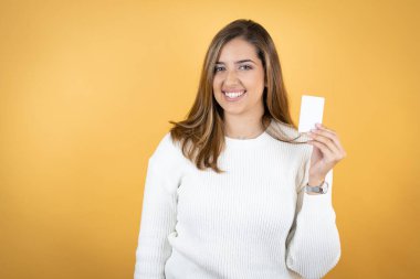 İzole edilmiş sarı arka planı olan beyaz bir kadın gülümsüyor ve elinde beyaz bir kart tutuyor.