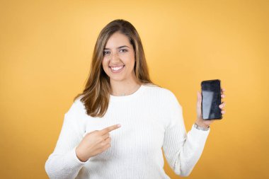 İzole edilmiş sarı arka planda genç beyaz kadın gülümsüyor ve telefonu işaret ediyor.