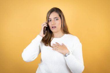 Kafkasyalı genç bir kadın izole edilmiş sarı arka planda telefonda endişeli bir ifadeyle konuşuyor.