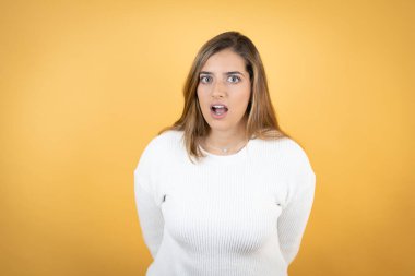 İzole edilmiş sarı arka planda genç beyaz kadın şaşkınlık ve şaşkınlık ifadesi, korku ve heyecan dolu yüz ifadesiyle korkmuş ve şok olmuştu..